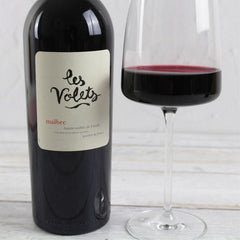 Bottle of Les Volets Malbec wine with a glass of red wine on a wooden surface.